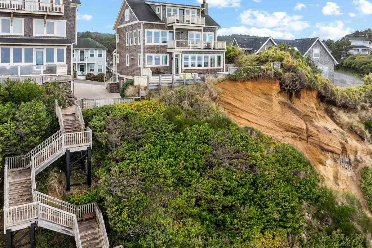 This image presents the multi-level house with a staircase leading down to the sandy beach, surrounded by lush cliffside vegetation. The view suggests easy access to Olivia Beach for walks at sunrise or sunset, reinforcing the connection between the property and its coastal surroundings.