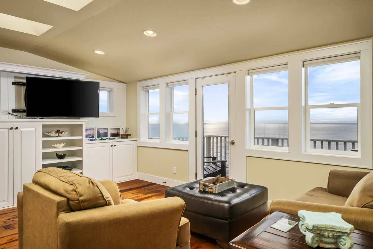 A comfortable seating area is arranged around a coffee table, with a large television unit positioned for easy viewing. Floor-to-ceiling windows showcase expansive ocean views, while soft natural light enhances the overall coziness. Plush sofas and a relaxed layout invite guests to unwind, read, or enjoy a movie together.