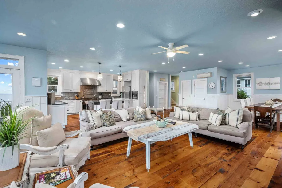 A spacious great room featuring a large sectional sofa, a coffee table with decorative items, and an open sightline to the kitchen area and dining space. The room is bathed in natural light, with a welcoming, comfortable atmosphere perfect for gatherings and relaxation.