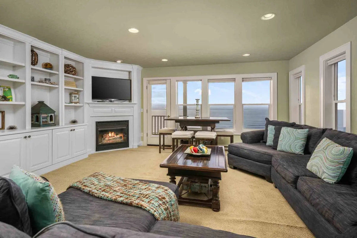 A light-filled interior with a sofa arranged near a modern fireplace, a dining table for group activities, and windows framing beautiful ocean vistas. The layout encourages both togetherness and moments of solitude, ideal for unwinding or entertaining.
