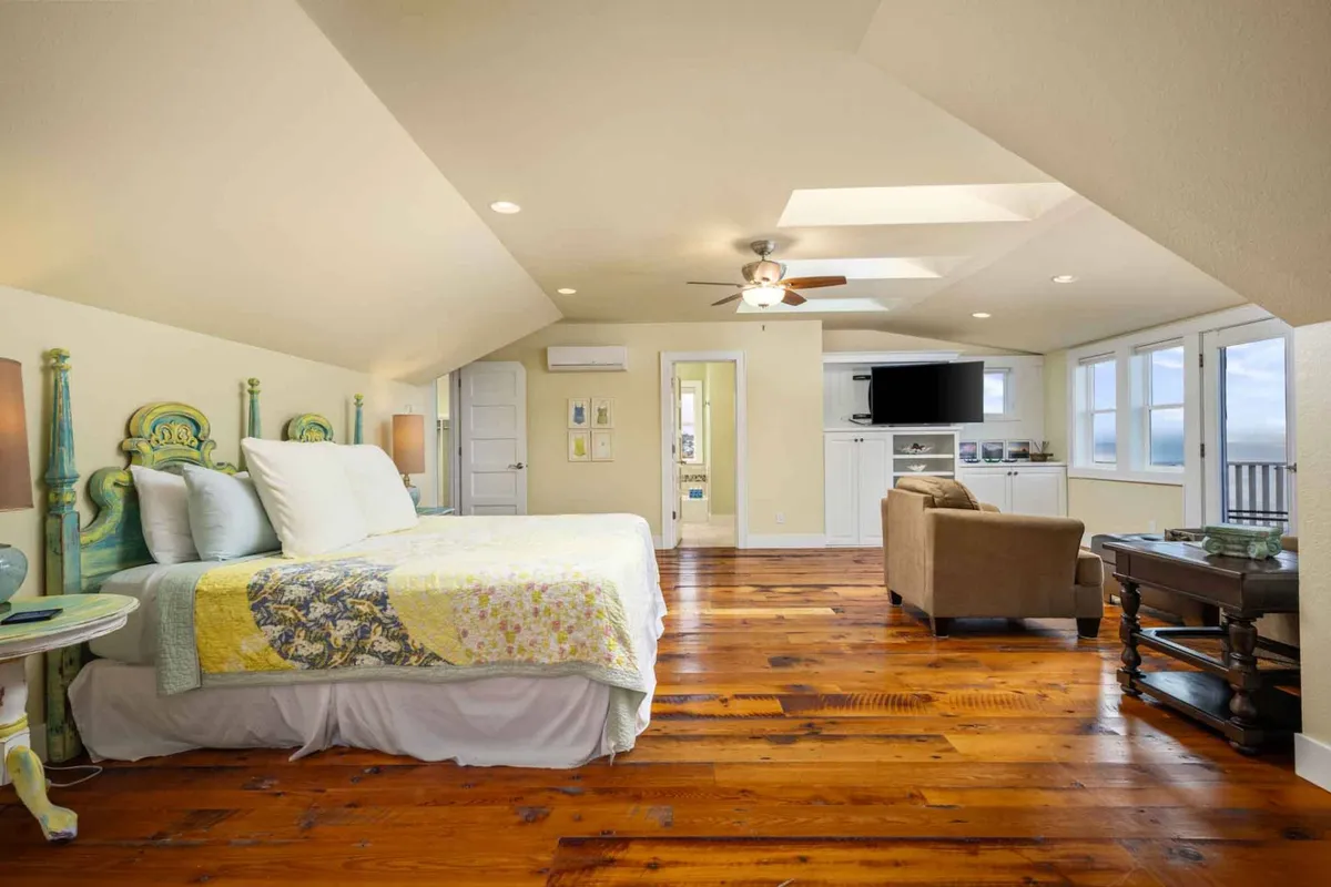 A tranquil bedroom featuring a decorative headboard, brightly colored quilt, wooden floors, and large windows allowing in natural light. A comfortable seating area adds to the relaxing ambiance.