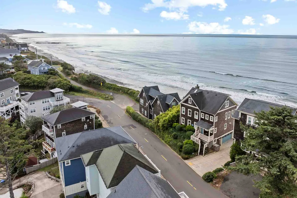 A wide, scenic view of the Oregon coastline, featuring the sparkling ocean, coastal homes, and a winding road that invites exploration. The open sky hints at seasonal changes, while the nearby beach suggests opportunities for walks, kite flying, and festival fun.