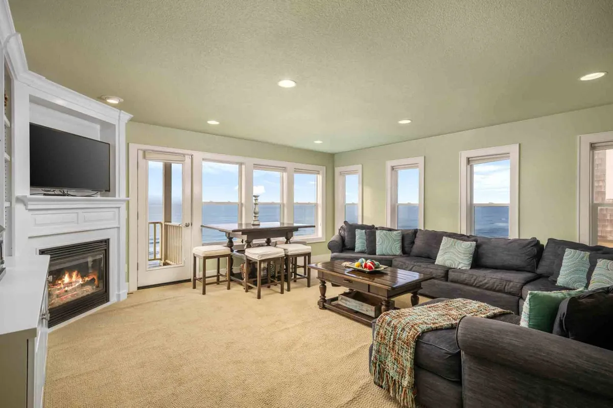 A cozy living and dining area with a large sectional sofa, a warm fireplace, expansive viewing windows offering ocean vistas, and a formal dining table. The setting exudes comfort and is perfectly suited for socializing, relaxing by the fire, or enjoying meals with a view.