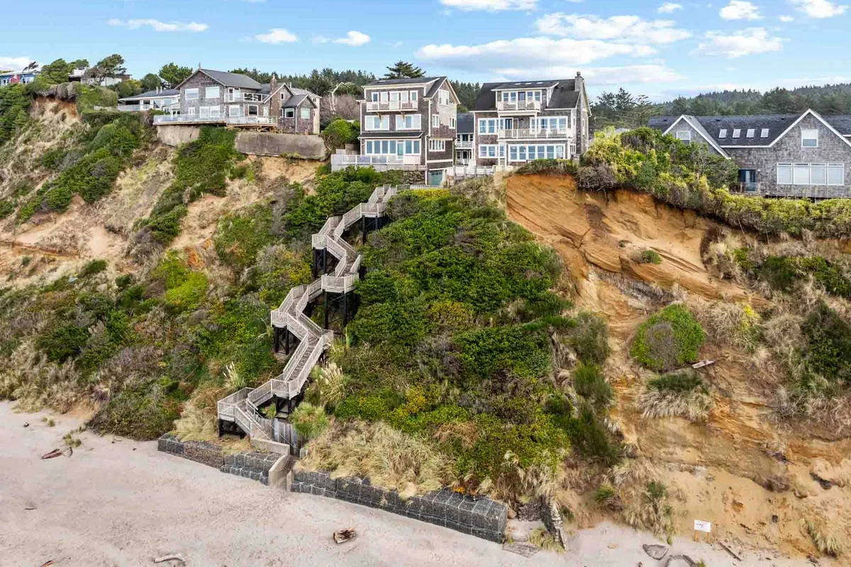A scenic view showcasing cliffside homes perched above a beautiful sand beach, with a winding staircase providing direct access down to the shoreline. Lush greenery surrounds the area, evoking a sense of adventure and family-friendly exploration.