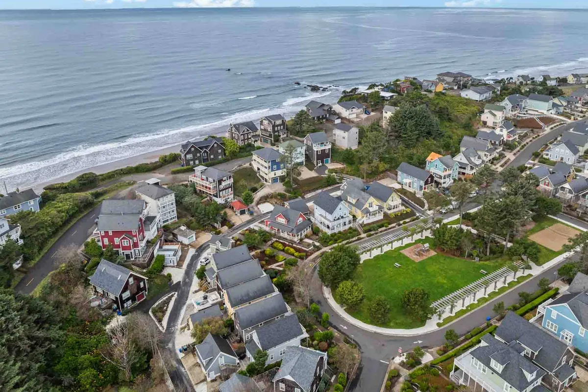 A broad view of the Olivia Beach community, showing the ocean, sandy beach, colorful coastal homes, a green park, and a tree line. The image highlights the communal atmosphere, access to outdoor recreation, and proximity to the shoreline.